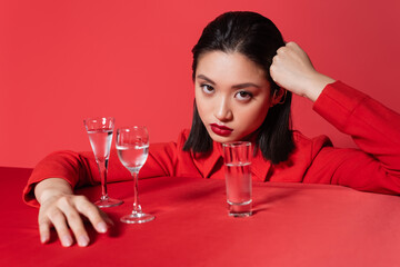 young asian woman in jacket holding hand near head and looking at camera near glasses with water isolated on red.