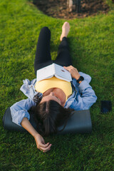 Top view of female in sporty clothes lying on green grass reading book, she take a deep breath on fresh air. Beautiful green. Fitness woman. Nature summer
