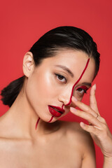 portrait of sensual asian woman with bright artistic makeup looking at camera and touching face isolated on red.