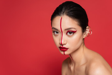 sexy asian woman with bare shoulders posing in artistic makeup and stylish ear cuff isolated on red.