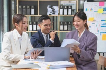 Team business three smiling and happy Asian men and women team using tablets and laptops brainstorm and plot statistics on a corporate chart board inside the company. Colleagues concept.