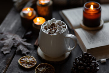 Autumn cozy home composition with hot chocolate with marshmallow and candles. Aromatherapy on a grey fall morning, atmosphere of cosiness and relax. Wooden background, books, close up.