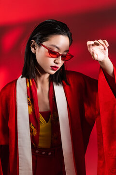 elegant asian woman in stylish sunglasses and kimono cape posing on red background with shadow.
