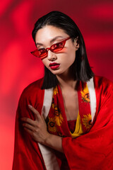 young asian woman in kimono cape and sunglasses looking at camera on abstract red background.