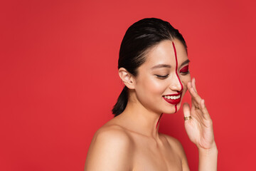 young asian woman with naked shoulders and artistic visage smiling with closed eyes isolated on red.