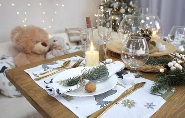 Christmas and New Year table setting with decorations. Festive dinner. A plate with a napkin, wine glasses, cutlery, candles and fir branches. Christmas tree and teddy bears in the background.