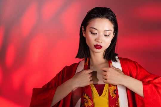 young asian woman in kimono cape touching neckerchief with floral print on abstract background with red gradient.