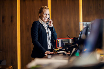 A happy receptionist is talking with guest on the phone and making the reservation while standing in hotel at reception.