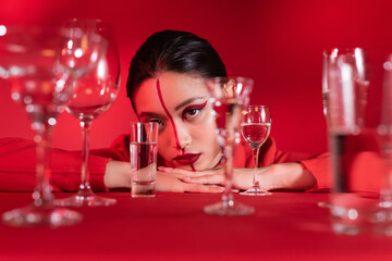 young asian woman with creative makeup on face divided with line near blurred glasses of water on red background.