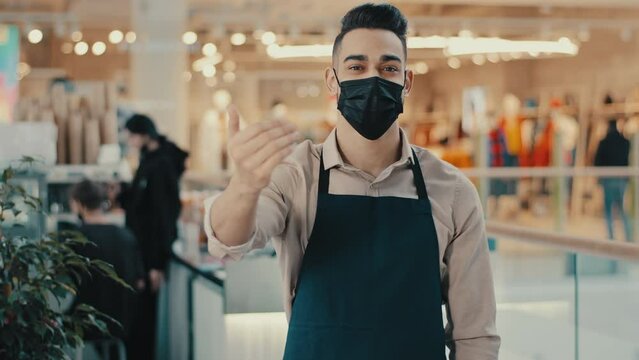 Friendly ethnic waiter guy cafe worker cafeteria restaurant shop manager small business owner in medical mask inviting welcoming salesman in apron come here invitation gesture welcome clients invite