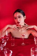 stylish asian woman with bare shoulders and artistic visage touching neck near various glasses with water on red background.