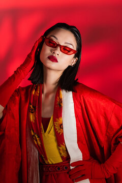 fashionable asian woman in kimono cape and sunglasses posing with closed eyes on red background with shadow.