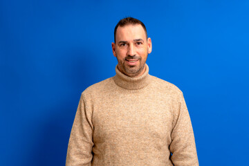 Fototapeta premium Isolated photo of handsome man with beard, mustache and trendy hairstyle, wears brown turtleneck sweater, has joyful expression while listening to his interlocutor, poses in studio blue background.