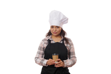 Isolated portrait on white background, of a multi-ethnic beautiful woman barista bartender, wearing chef's hat and black apron, posing with takeaway coffee in disposable paper cup, looking at camera