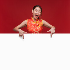 Young asian woman wearing qipao cheongsam dress with hand point to copy space on red background for...