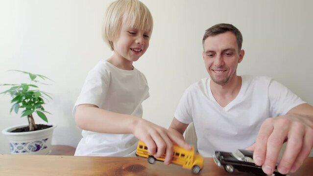 Son and dad are playing with toy cars, smiling and laughing at the desk against the white wall. Happy father and son are having a great time together at home