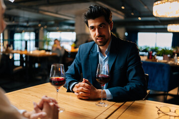 View from back of unrecognizable young woman to smiling elegant man in suit sitting at table with glasses of red wine having festive dinner at luxury restaurant. Happy loving couple enjoying talking.