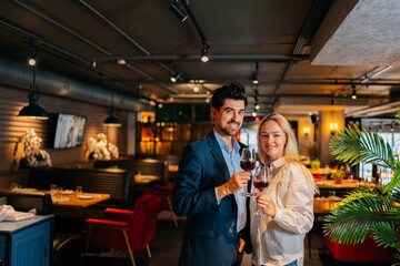 Portrait of positive elegant man and woman holding glasses of wine, looking at camera, standing in fancy restaurant at evening. Happy loving couple enjoying romantic dinner, celebrating anniversary.