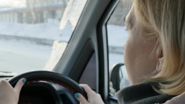The driver is an adult blonde woman driving on a winter snowy road driving a car with a right-hand drive. The woman enjoys driving.