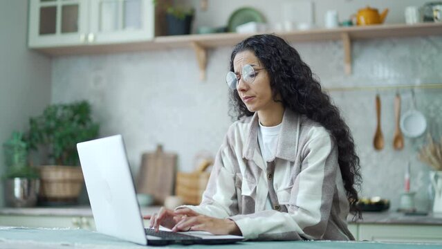 Curly woman with glasses sit in the light kitchen feels annoyed having problems with laptop computer Upset girl can't work because system error data loss backup battery issues forgot password at home 
