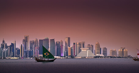 Obraz premium Traditional boat sail with Brasil country flag design for FIFA World Cup 2022 in Corniche area with amazing sunset view of cityscape skyscraper buildings in Doha, Qatar