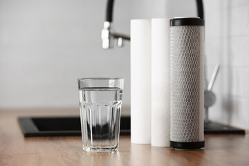 A glass of clean fresh water and set of filter cartridges on wooden table in a kitchen interior. Installation of reverse osmosis water purification system. Concept Household filtration system.