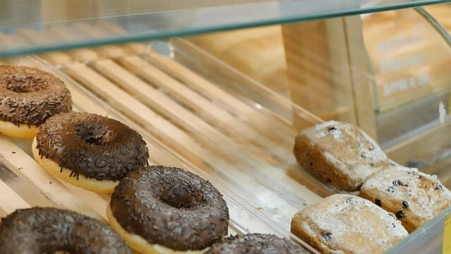 A customer picks up sweet pastries, cupcakes, and donuts from a display case in a bakery using serving tongs. Baking and flour food threatens to increase excess weight.