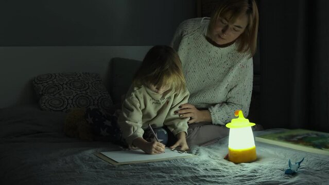 Mother and little daughter studying and drawing in a complete darkness during electricity outage. Little girl uses camping lantern to do her homework during blackout. Energy crisis concept