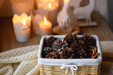 Basket with pine cones and seasonal spices on the table. Lit candles in the background. Hygge at home. Selective focus.