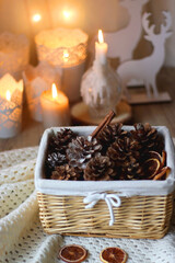 Basket with pine cones and seasonal spices on the table. Lit candles in the background. Hygge at home. Selective focus.