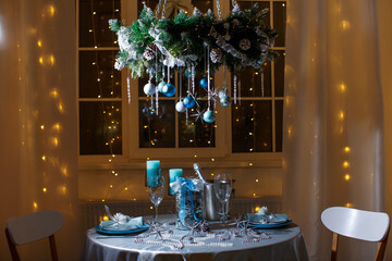 A Christmas table for two with candles, glasses, a bottle of sparkling wine against the backdrop of a night window with garland lights. Chandelier on chains, with balls, stars, cones and icicles