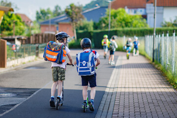 Two school kid boys in safety helmet riding with scooter in the city with backpack on sunny day. Happy children in colorful clothes biking on way to school. Safe way for kids outdoors to school