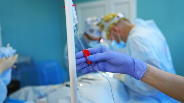 Female hand in latex blue glove touching the roller at drop counter. Two surgeons bent over the patient performing surgery at backdrop in blur.