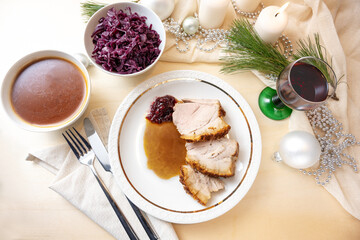 Roast pork slices with crust on a plate served on a festive table with sauce, red cabbage, wine glass, white candles and Christmas decoration, holiday dinner, top view from above