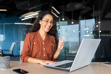 Plakat Cheerful and successful Latin American businesswoman talking remotely via video call with colleagues and partners, office worker inside the office using a laptop at work.
