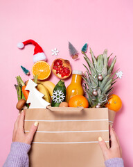 Woman holding paper bag with Christmas food for donation on pink background