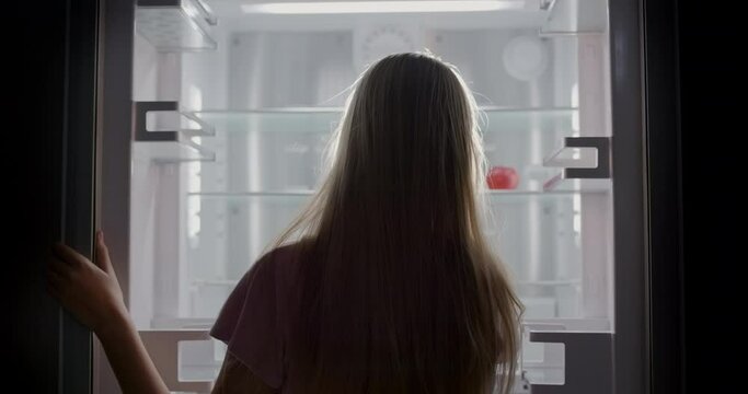 A young girl looks into an empty refrigerator where one red apple lies. Diet and weight loss concept