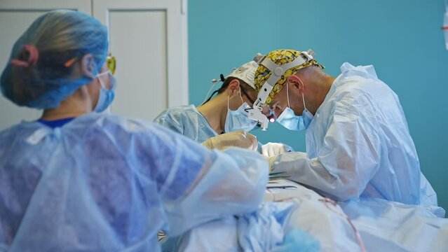 Male doctors in caps with funny pictures and device flashlights on their heads collaborate at surgery. Female nurse standing her back to camera prepares instruments for the specialists.