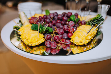 A variety of fruits, grapes, pineapple lie on a mirrored tray on a table with skewers in a restaurant at a wedding. Food photography, banquet, buffet.