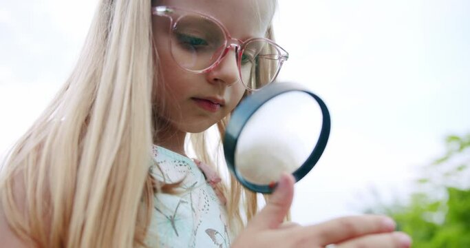 Cute curious girl with magnifying glass in nature. Low angle handheld shot of little blond girl in big glasses exploring bug on finger looking through big magnifying glass in summer park