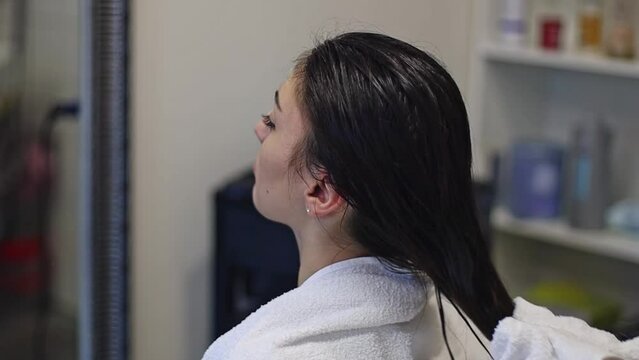 Beautiful young caucasian girl hairdresser dries wet long brown hair with a white towel to a client sitting in a chair in a hairdressing salon, side view close-up and slow motion.