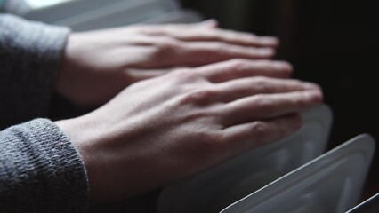 Woman warms hands from electric heater oil radiator indoors. Close-up shot. Cold winter, economic crisis concept. Comfort, winter heating and heat conservation. 