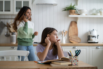 Thoughtful unconcentrated girl look to side doing homework sits at table with textbooks. Woman talking on mobile phone standing in kitchen behind school-age daughter doing extracurricular work