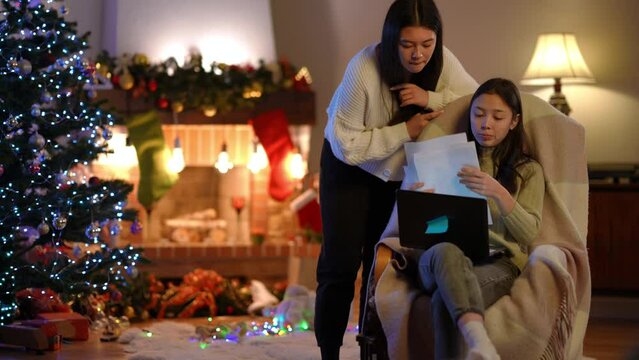 Two focused Asian young women analyzing paperwork sitting with laptop in living room on New Year's eve. Concentrated freelancers planning business on Christmas indoors on the right. Slow motion