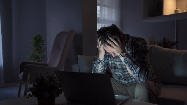Man working from home looking at laptop and seeing something bothering him and getting upset.
Man working remotely from home at night doing his work on laptop over internet feeling upset and stressed.