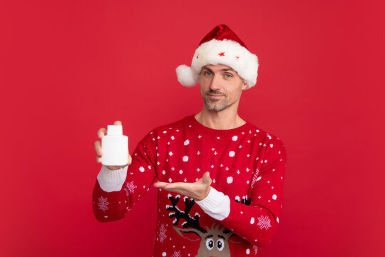 Man in Christmas sweater and hat hold bottle of medicine on color background. Middle aged santa.