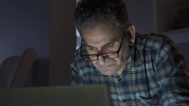 Mature man looking at laptop at night at home doing serious work and focusing.
Focused and serious man looking at laptop at home.
