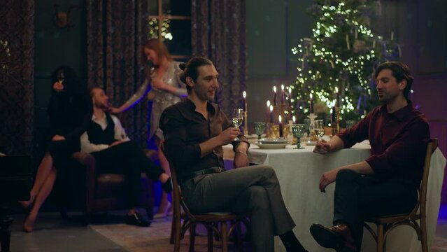 Two very handsome and good looking men very sensual discussing together while celebrating the Christmas they sitting together at the dining table on the background their friends discussing while