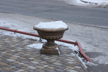 one brown concrete flowerpot stands on the sidewalk in white snow on a winter street near a gray asphalt road