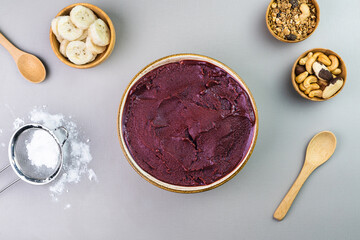 Brazilian açai in a white bowl, additional banana, granola and nuts in small bamboo bowls. Cashew and Pará nuts. Top view. Gray background. Selective focus